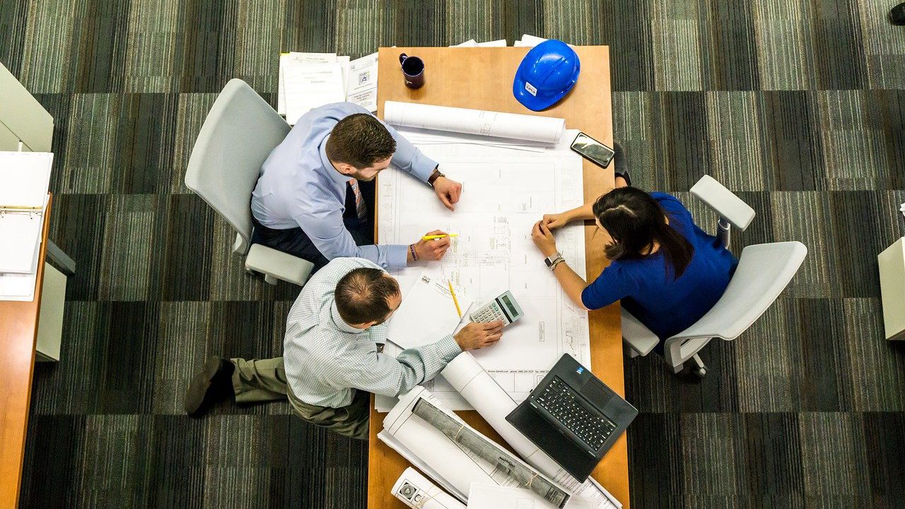 Three office workers sitting around a table with blueprints at a desk.